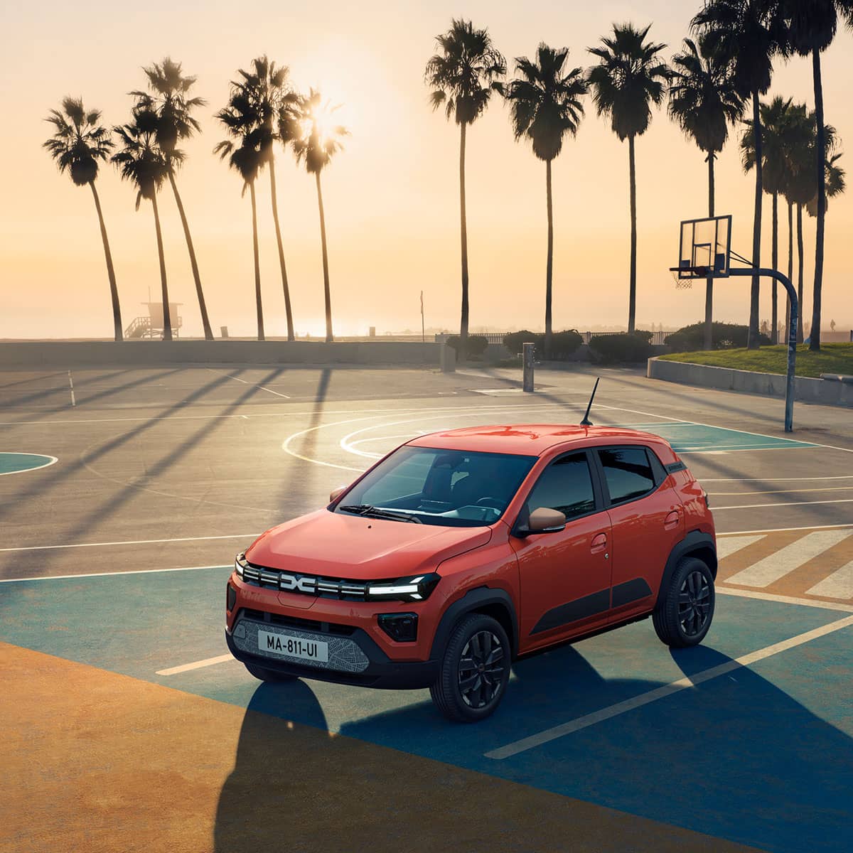 Dacia Spring im Sonnenuntergang auf einem Basketballfeld vor Palmen